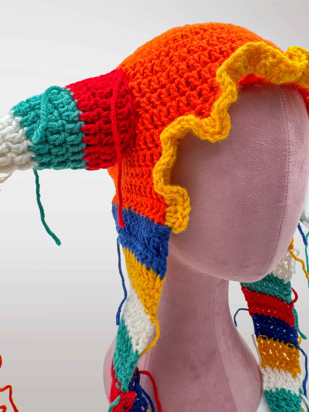 A colorful crocheted jester-style hat features an red-orange crown, yellow ruffled trim, and multicolored striped side pieces with tassels and long dangling yarn tails.