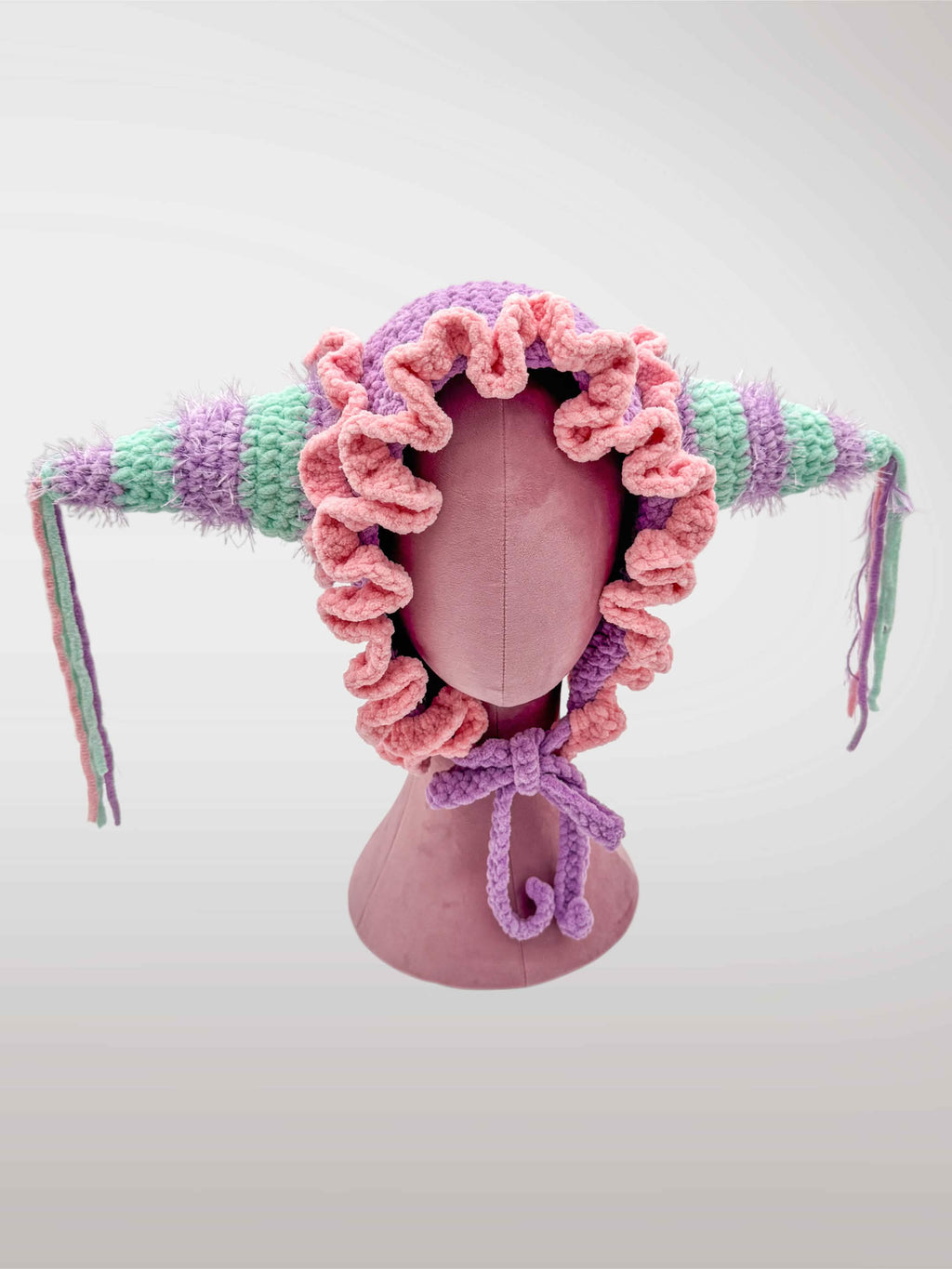 A handmade crocheted jester-style bonnet with ruffles in pastel pink and purple, featuring jester points with dangling tassels. 