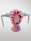 A handmade crocheted jester-style bonnet with ruffles in pastel pink and purple, featuring jester points with dangling tassels. 