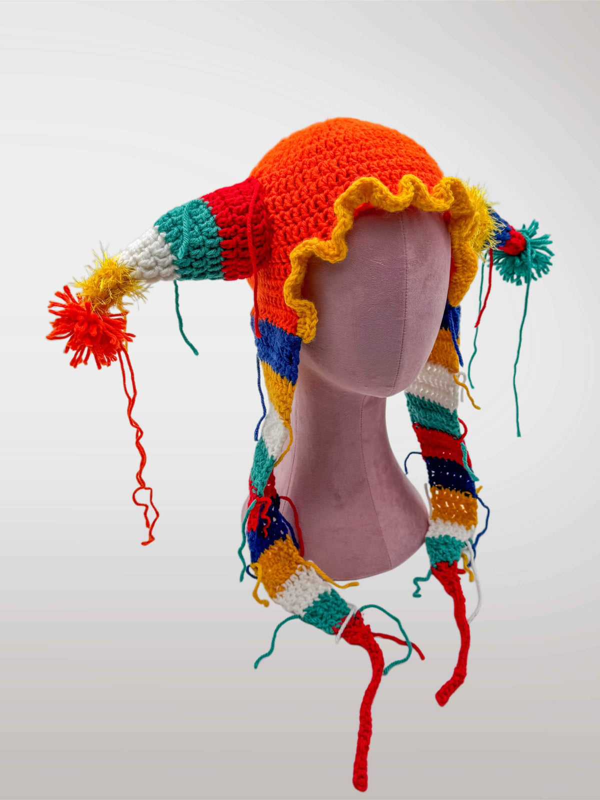 A colorful crocheted jester-style hat features an red-orange crown, yellow ruffled trim, and multicolored striped side pieces with tassels and long dangling yarn tails.