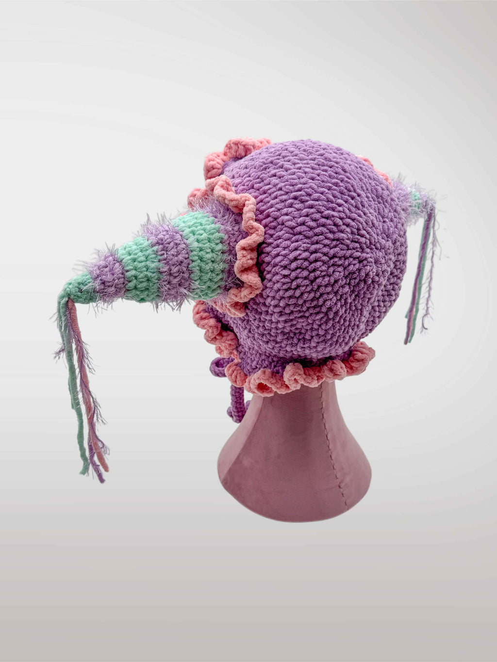 A handmade crocheted jester-style bonnet with ruffles in pastel pink and purple, featuring jester points with dangling tassels. 