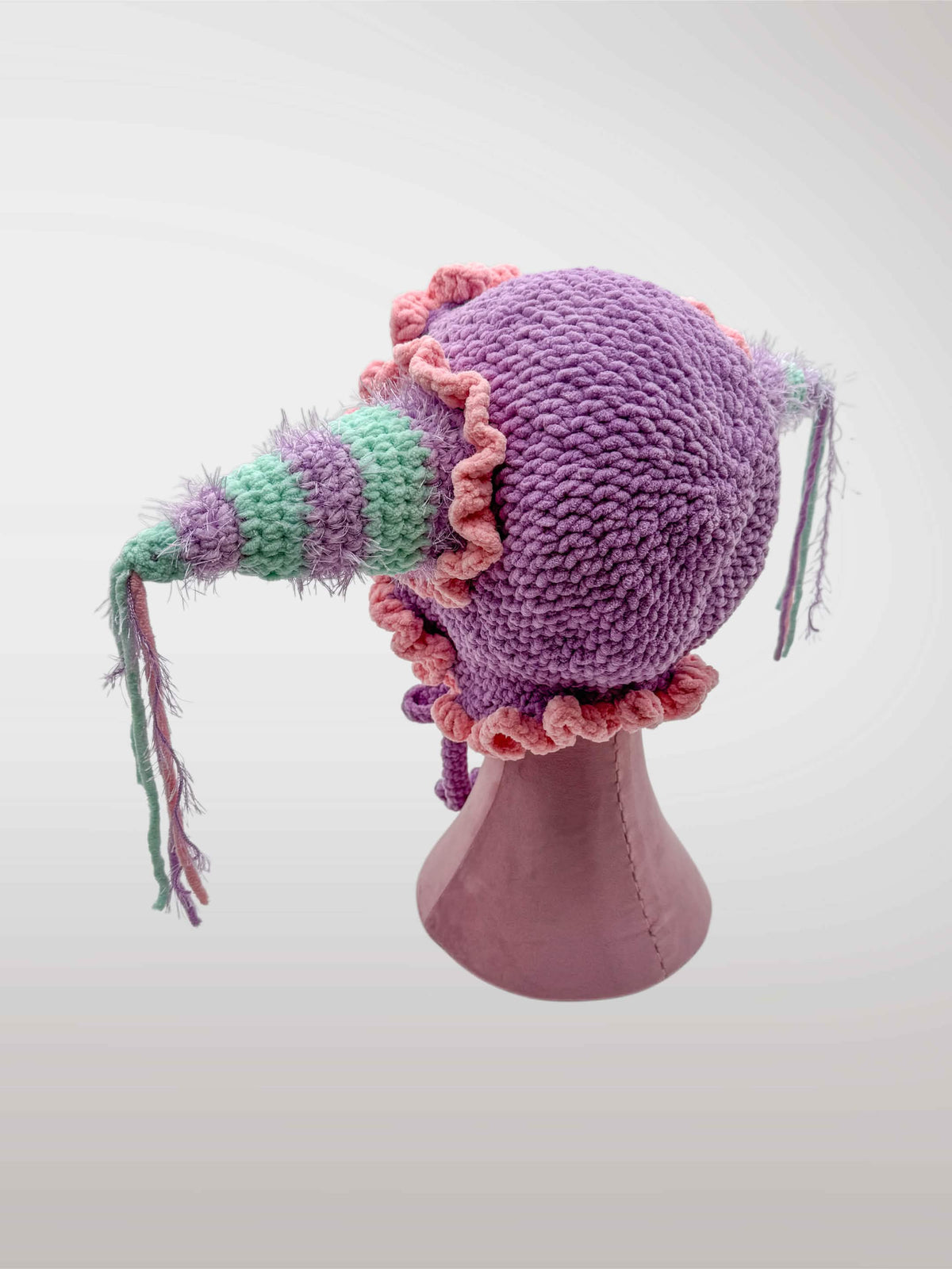 A handmade crocheted jester-style bonnet with ruffles in pastel pink and purple, featuring jester points with dangling tassels. 