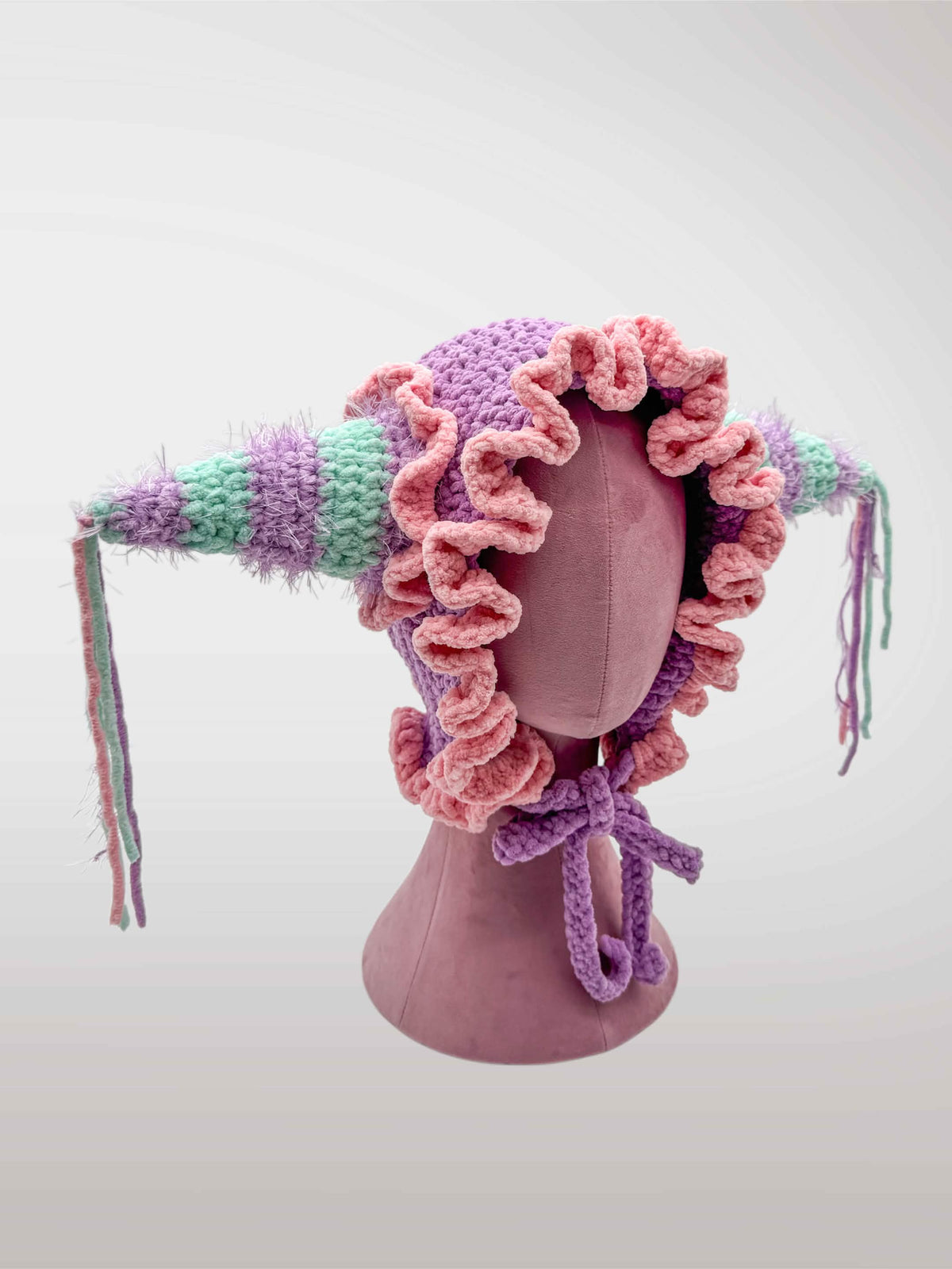 A handmade crocheted jester-style bonnet with ruffles in pastel pink and purple, featuring jester points with dangling tassels. 
