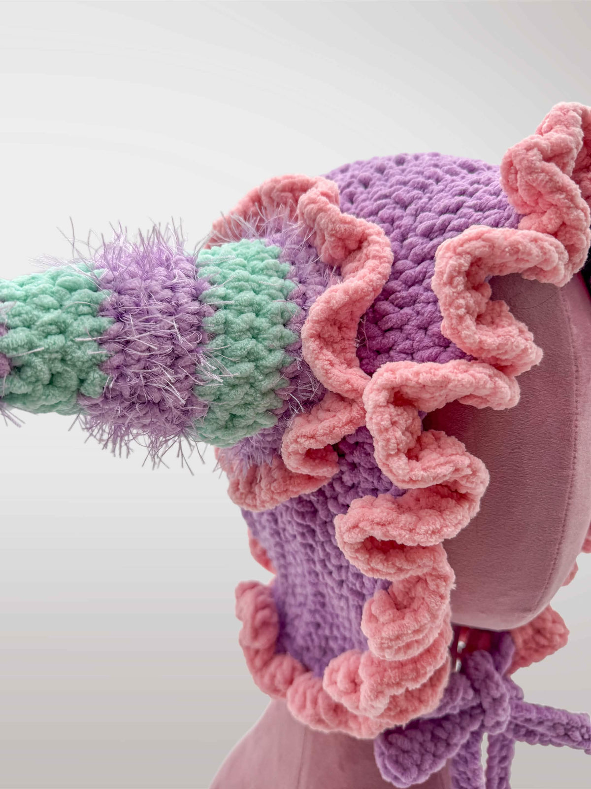 A handmade crocheted jester-style bonnet with ruffles in pastel pink and purple, featuring jester points with dangling tassels. 