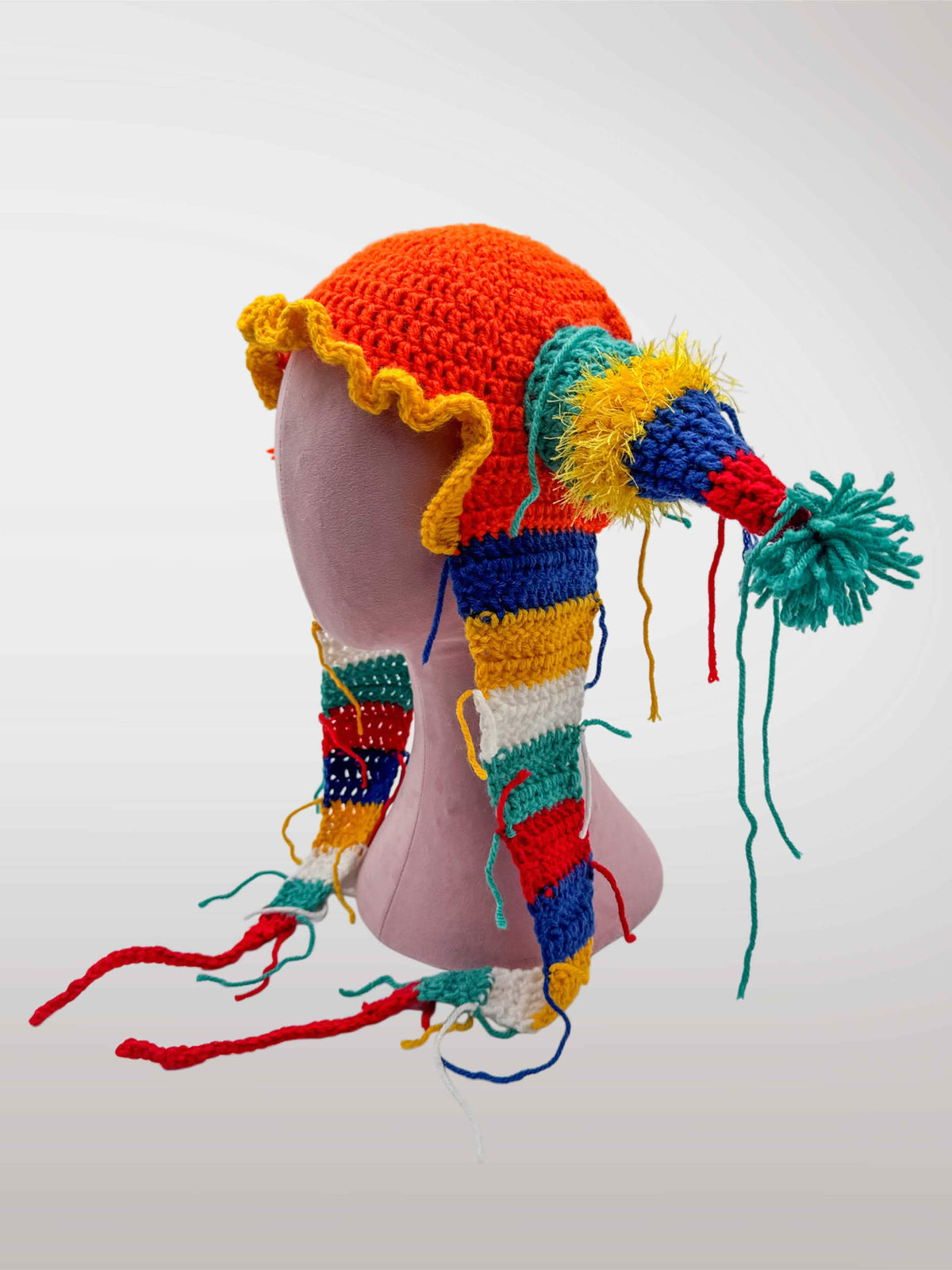 A colorful crocheted jester-style hat features an red-orange crown, yellow ruffled trim, and multicolored striped side pieces with tassels and long dangling yarn tails.