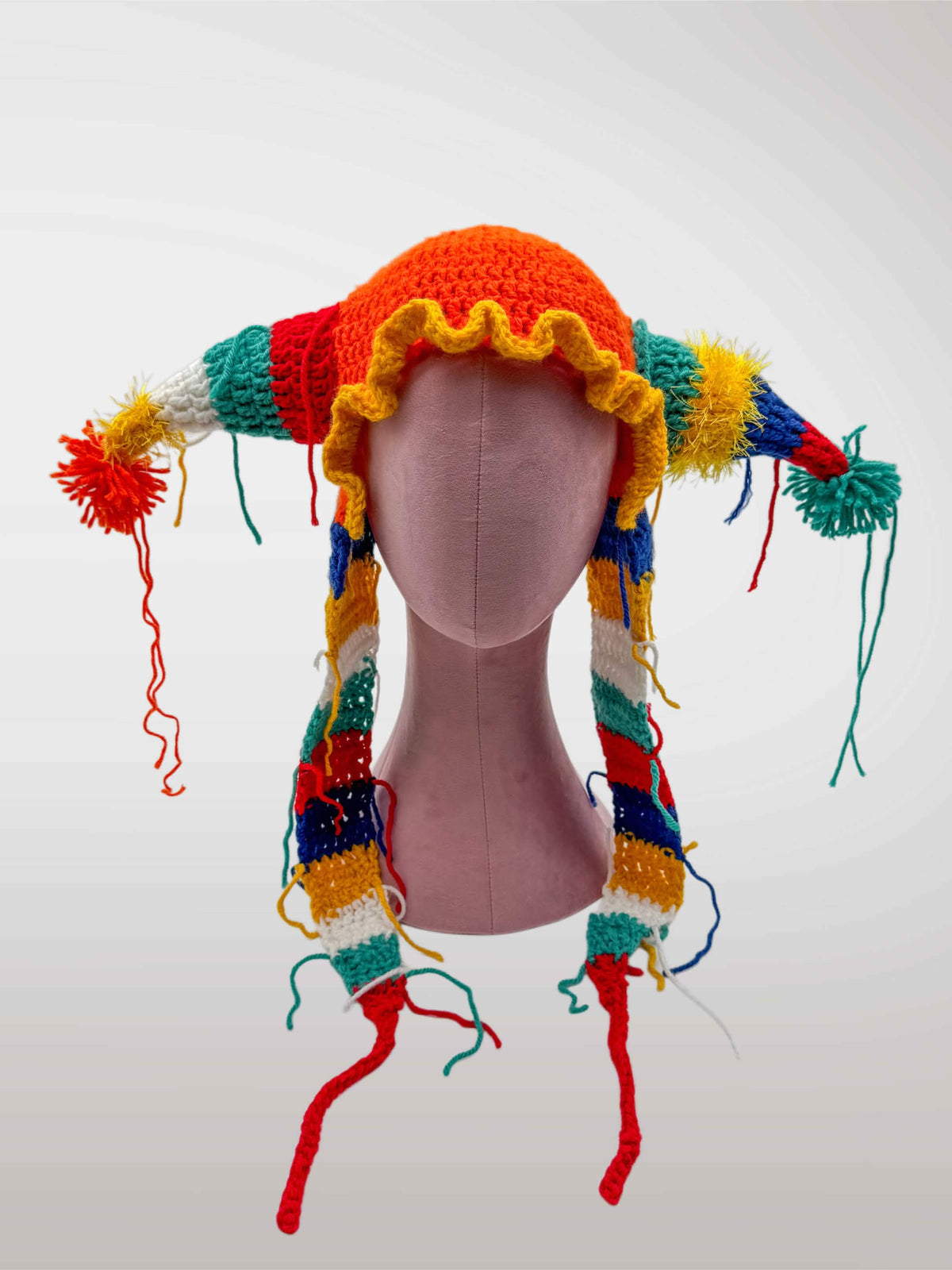A colorful crocheted jester-style hat features an red-orange crown, yellow ruffled trim, and multicolored striped side pieces with tassels and long dangling yarn tails.